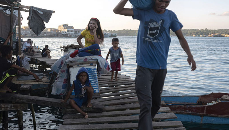 A father carrying a bag while one kid helps, another kid is sitting on the ground, and the wife is on a call.